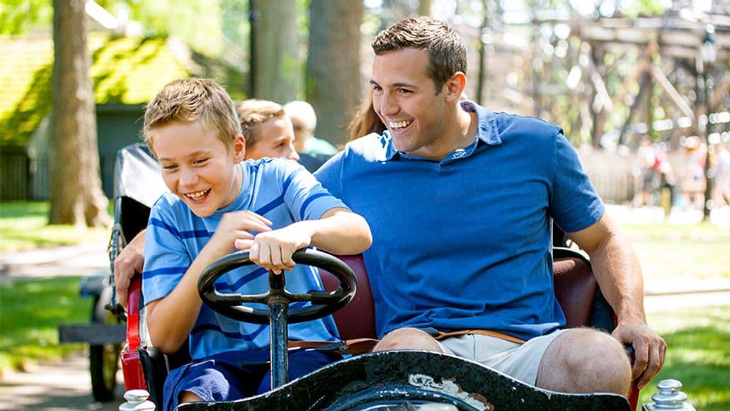 A father and son driving an antique car at an amusement park.